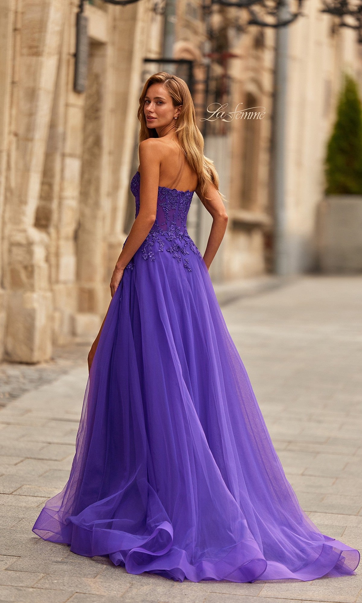 A woman in the La Femme 33551 long prom dress, featuring floral embroidery and a sheer corset bodice, stands on a cobblestone street looking over her shoulder.
